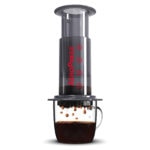 Badger & Dodo. The Aeropress Starter Kit brews rich coffee into a glass mug, droplets falling from the press—an ideal choice for any coffee lover.