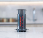 Badger & Dodo. A black Aeropress Starter Kit rests on a marble countertop with a blurred kitchen background—perfect for brewing your next cup.