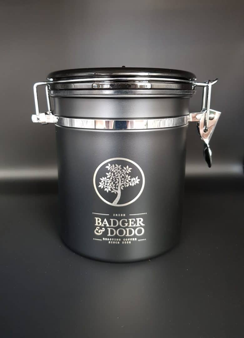 Badger and Dodo Coffee Roasters: A black metal coffee canister with a silver latch and airtight seal, featuring Badger & Dodo roastery branding and a tree logo, set against a plain dark background.