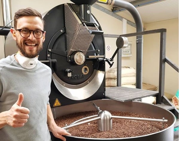 Badger and Dodo Coffee Roasters: A person with glasses gives a thumbs-up next to a large coffee roasting machine filled with future crops of coffee beans. They are wearing a grey shirt and a mask around their neck. Bags of coffee beans are stacked in the background.