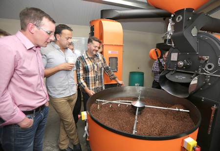 Badger and Dodo Coffee Roasters: A group of men stand around an industrial coffee roasterys machine filled with brown beans. One man holds a cup, and they appear to be observing the roasting process. The large, orange machine features prominent control panels and dials, marking an insightful visit to the facility.