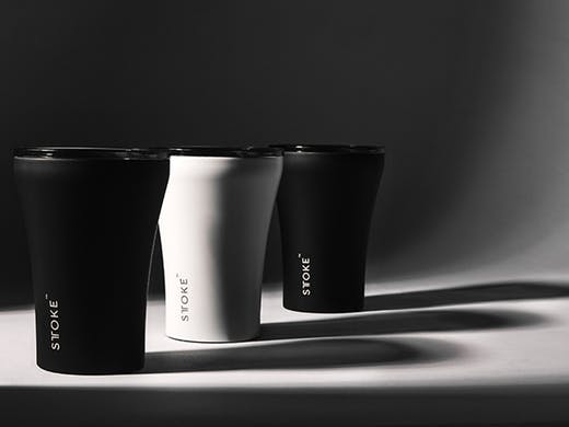 Badger and Dodo Coffee Roasters: Three insulated reusable cups labeled STŌKE are aligned on a surface. Two are black, and one is white, casting long shadows behind them. The minimalist, high-contrast effect hints at an expanding range that blends style with sustainability.