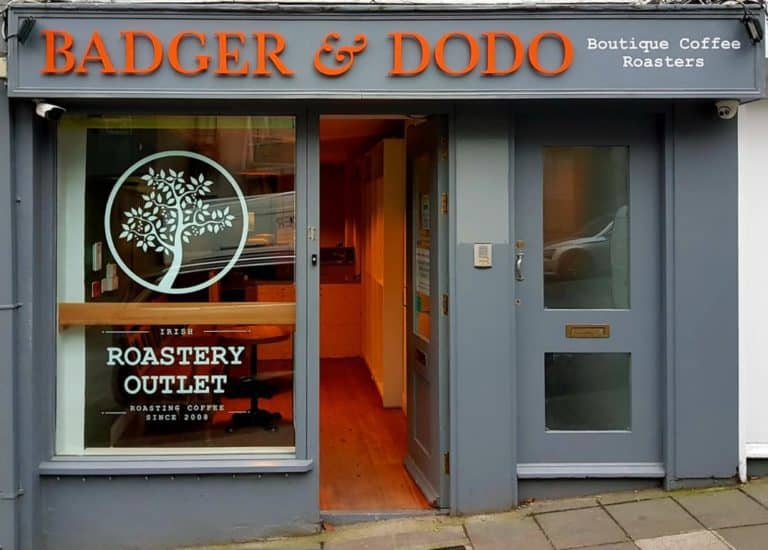 Badger and Dodo Coffee Roasters: The storefront of Badger & Dodo, a boutique coffee roaster, boasts gray walls adorned with vibrant orange and white lettering. A sign proclaims it as an Irish Roastery Outlet alongside a tree logo, marking this retail roasterys charming facade on the bustling sidewalk.