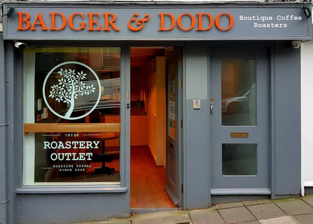 Badger and Dodo Coffee Roasters: The storefront of Badger & Dodo, a boutique coffee roaster, boasts gray walls adorned with vibrant orange and white lettering. A sign proclaims it as an Irish Roastery Outlet alongside a tree logo, marking this retail roasterys charming facade on the bustling sidewalk.