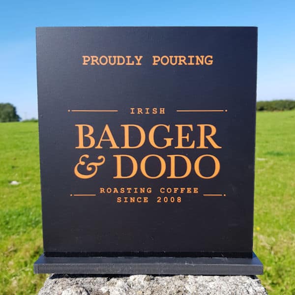 Badger and Dodo Coffee Roasters: A shelf sign stands outdoors, with bold orange letters reading: Proudly Pouring Irish Badger & Dodo Roasting Coffee Since 2008 on a sleek black background. The vibrant colors pop against the lush green grass and clear blue sky.