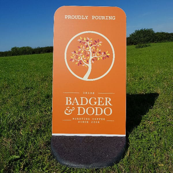 Badger and Dodo Coffee Roasters: An Ecoflex pavement sign on the grass reads, Proudly Pouring Irish Badger & Dodo Roasting Coffee Since 2008. The orange sign, adorned with a tree graphic, stands out beautifully against the clear blue sky.