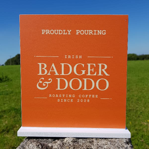 Badger and Dodo Coffee Roasters: Against a grassy landscape and clear blue sky, an orange shelf sign reads, Proudly Pouring Irish Badger & Dodo Roasting Coffee Since 2008.