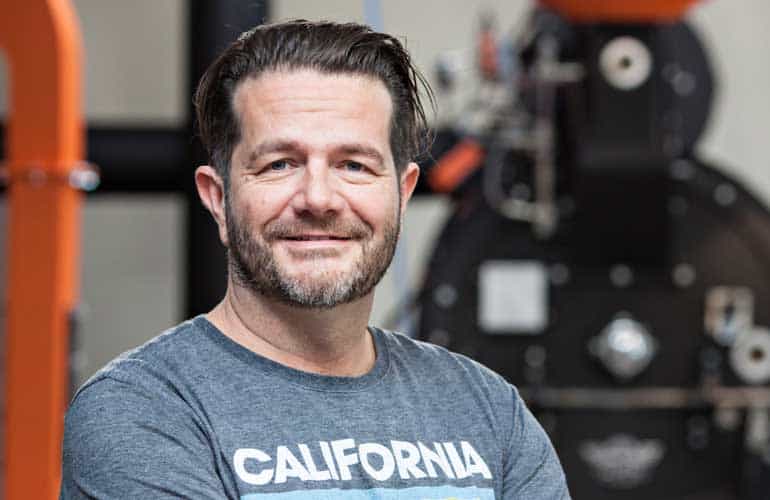 Badger and Dodo Coffee Roasters: Man with a beard and mustache smiling, wearing a gray California t-shirt, with arms crossed. He stands in a setting with industrial equipment, featuring orange and black colors in the background.