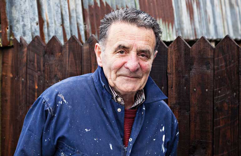 Badger and Dodo Coffee Roasters: Elderly man in a blue paint-splattered jacket stands in front of a wooden fence and metal roofing. He has short, gray hair and a gentle expression.