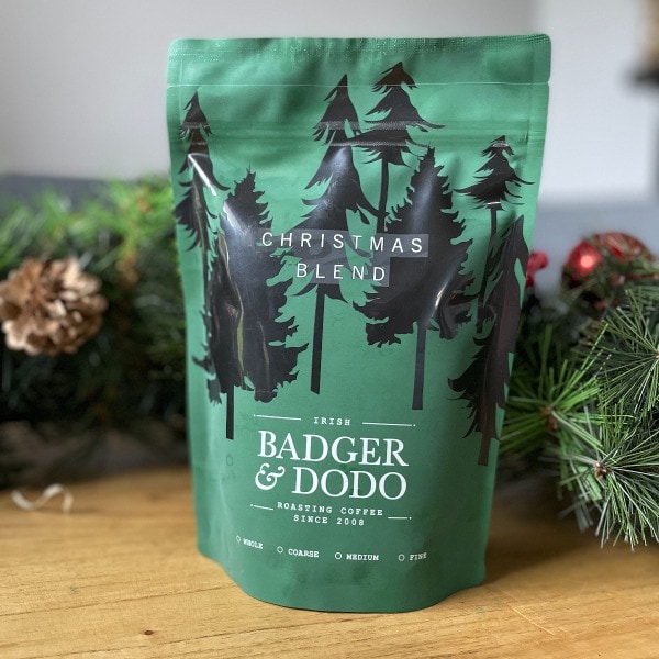 Badger and Dodo Coffee Roasters: A green coffee package labeled Christmas Blend from Badger & Dodo, adorned with festive pine trees, rests on a wooden surface. Surrounding it are holiday decorations like pine cones and greenery, all softly blurred in the background to celebrate this seasonal roast.