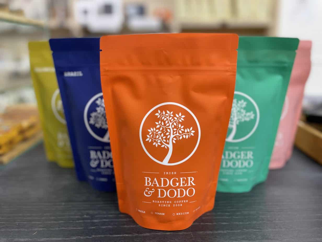 Badger and Dodo Coffee Roasters: Multiple colorful coffee bags are artfully displayed on a wooden table. The closest, an orange bag with a white tree logo, proudly reads Badger & Dodo Roasting Coffee Since 2008. These sustainable bags serve as vibrant companions for your coffee journey. In the background, blue, green, and yellow join the scene.