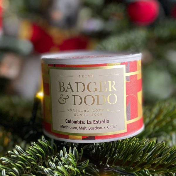 Badger and Dodo Coffee Roasters: A tin of Badger & Dodo coffee, labeled Colombia: La Estrella, rests on a green fir branch. The label notes flavors of mushroom, malt, Bordeaux, and cedar, set against a blurred festive background.