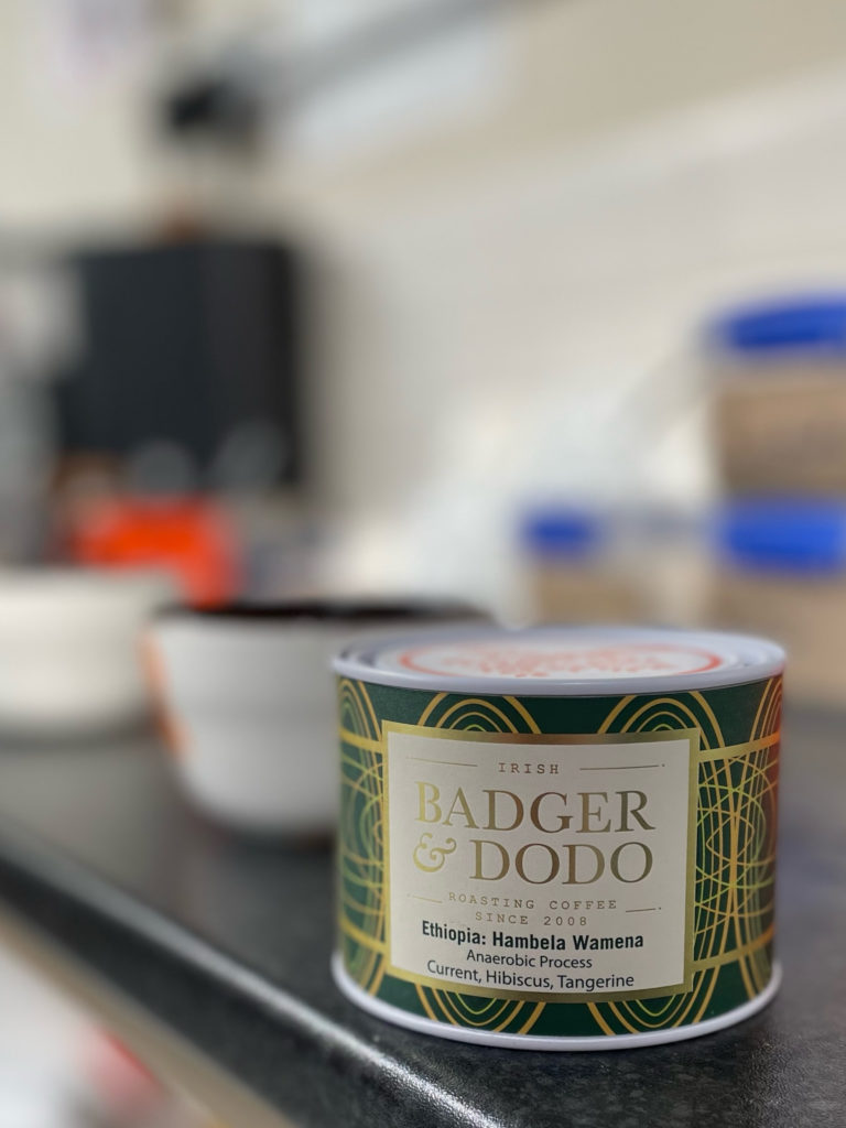 Badger and Dodo Coffee Roasters: A close-up of a Badger & Dodo coffee container highlights the Limited Edition Ethiopian Hambela Wamena with anaerobic processing. The can boasts notes of currant, hibiscus, and tangerine, set against a backdrop of blurry kitchen items.