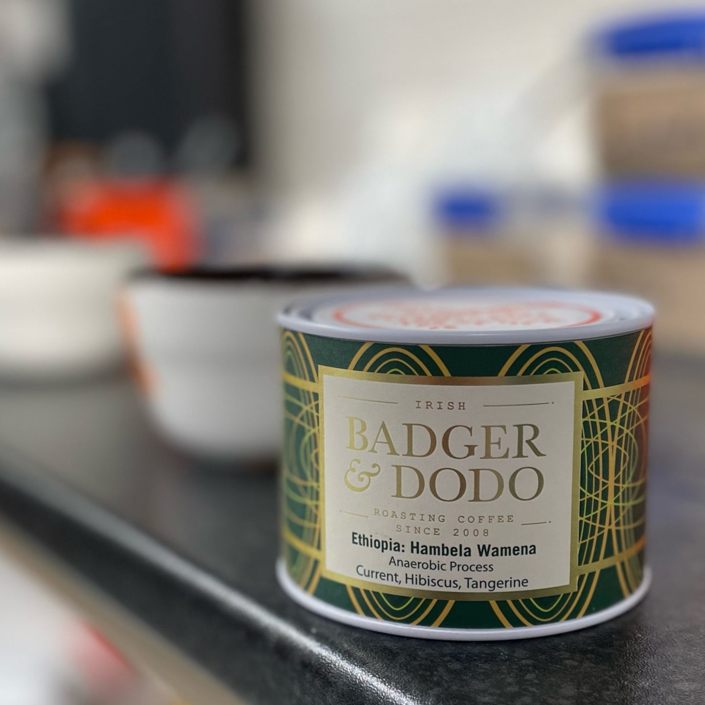 Badger and Dodo Coffee Roasters: A close-up of a Badger & Dodo coffee container highlights the Limited Edition Ethiopian Hambela Wamena with anaerobic processing. The can boasts notes of currant, hibiscus, and tangerine, set against a backdrop of blurry kitchen items.