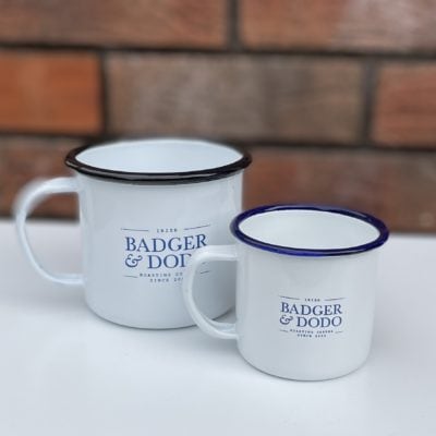 Badger and Dodo Coffee Roasters: Two Badger & Dodo Vintage Enamel Mugs with blue rims, featuring the logo in blue text. The larger mug is on the left, and the smaller on the right, placed elegantly on a white surface against a brick wall backdrop.