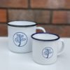 Badger and Dodo Coffee Roasters: Two Badger & Dodo Vintage Enamel Mugs, featuring white enamel with blue rims and tree designs, rest on a white surface against a blurred brick wall backdrop. A larger mug with a black rim adds contrast to the enamel set.