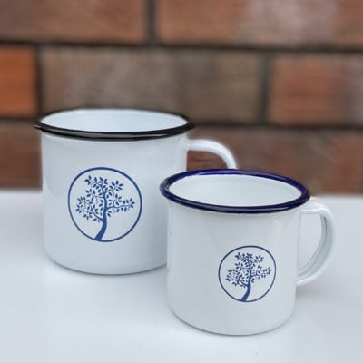 Badger and Dodo Coffee Roasters: Two Badger & Dodo Vintage Enamel Mugs, featuring white enamel with blue rims and tree designs, rest on a white surface against a blurred brick wall backdrop. A larger mug with a black rim adds contrast to the enamel set.