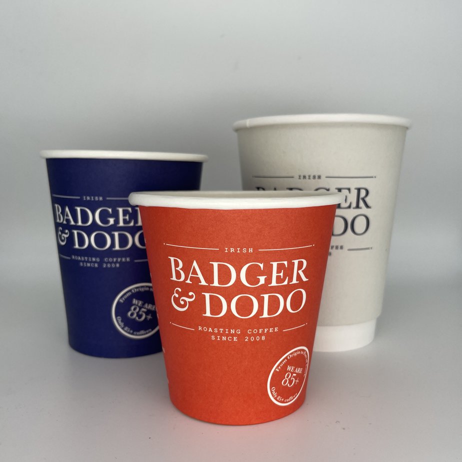 Badger and Dodo Coffee Roasters: Three disposable coffee cups with the Badger & Dodo logo shine in hues of red, blue, and white. Each cup boasts text about Irish Roasting Coffee Since 2008. Perfectly arranged against a plain background, these cups could grace any coffee wholesale catalogue.
