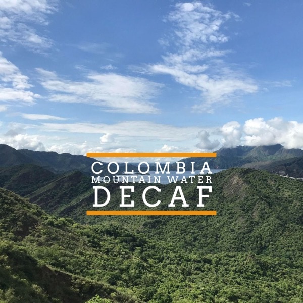 Badger and Dodo Coffee Roasters: Mountain range with lush greenery under a blue sky with clouds. Overlaid text reads Colombia Mountain Water Decaf in bold white and orange lettering.