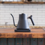 Badger & Dodo. The FELLOW STAGG EKG Electric Pouring Kettle in matte black with a long spout sits on a wooden kitchen counter.