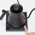 Badger & Dodo. A hand presses a button on the FELLOW STAGG EKG Electric Pouring Kettle in matte black, featuring a gooseneck spout and digital temperature display.