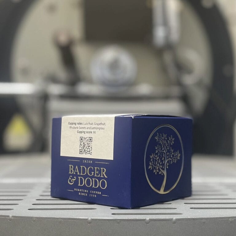 Badger & Dodo. A blue COLOMBIA–Jairo Arcila, Anaerobic coffee box from Badger & Dodo rests on a metal surface, evoking rich Colombian flavor, with a blurred machine in the background.