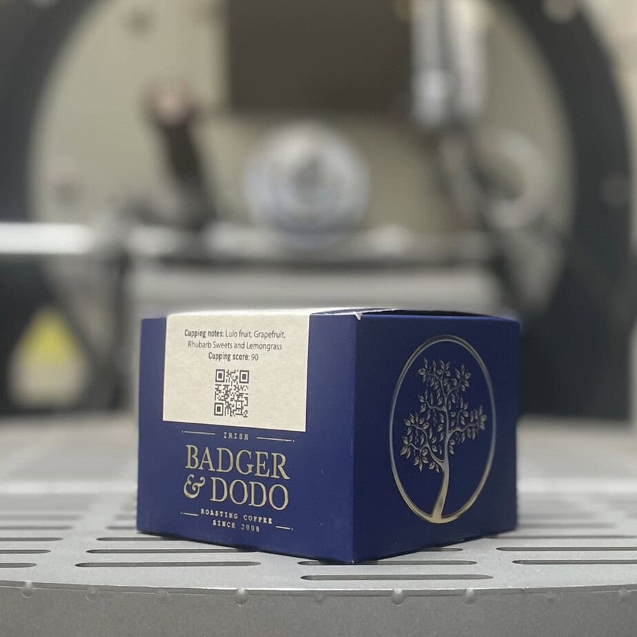 Badger & Dodo. A blue COLOMBIA–Jairo Arcila, Anaerobic coffee box from Badger & Dodo rests on a metal surface, evoking rich Colombian flavor, with a blurred machine in the background.