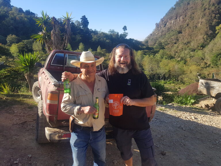 Badger & Dodo. Two men smiling, holding drinks, stand by a red pickup truck in a scenic, hilly outdoor area—capturing the spirit of an origin trip Guatemala adventure.