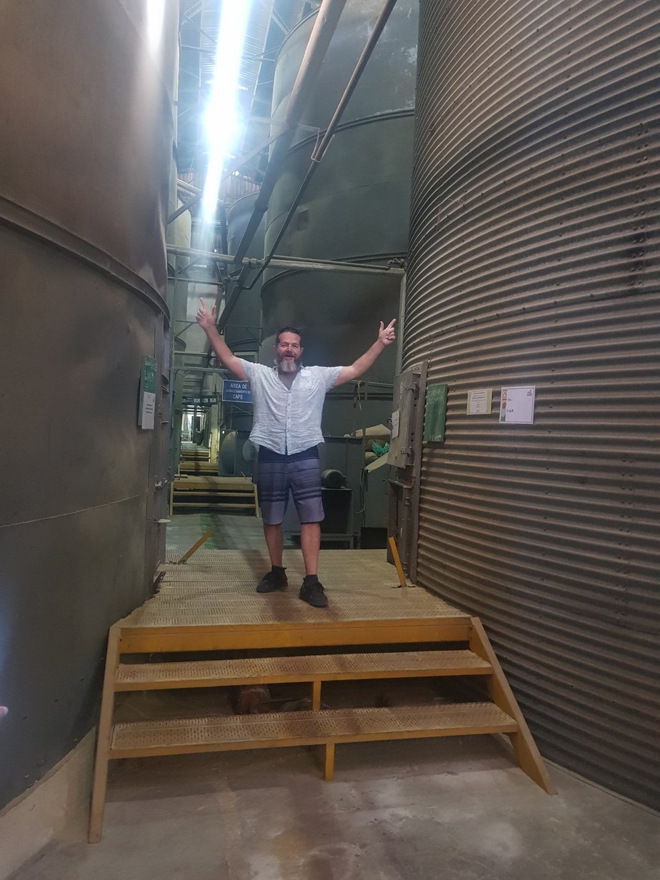 Badger & Dodo. A man stands with arms raised on metal stairs between large industrial storage tanks indoors, capturing the excitement of an origin trip in Costa Rica.