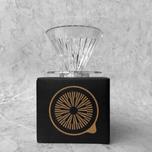 Badger & Dodo. The Filter V01 Glass - Crystal Eye with a white holder by Timemore rests on a sleek black scale featuring a gold circular pattern, all set against a marble background.