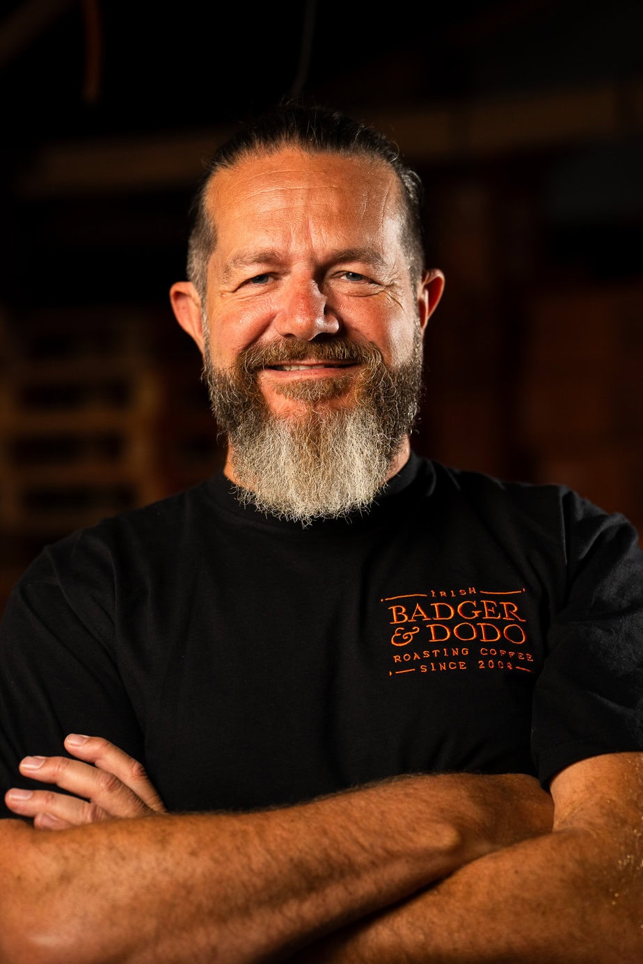 Badger & Dodo. Meet The Team: Smiling bearded man with arms crossed, wearing a black Badger & Dodo t-shirt, stands indoors.