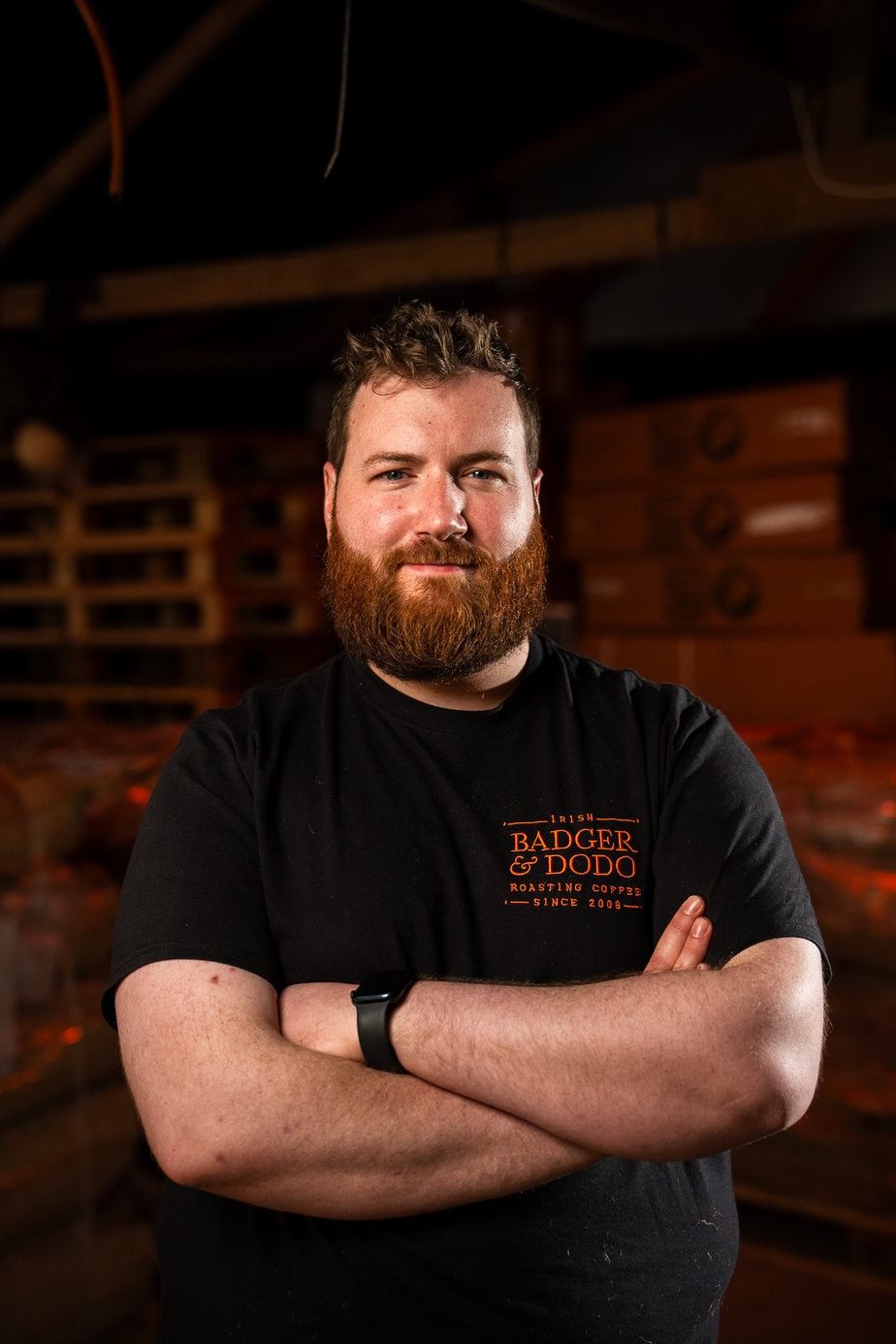 Badger & Dodo. Bearded man in a black t-shirt stands with arms crossed in a warm, dimly lit room—ready for you to meet the team.