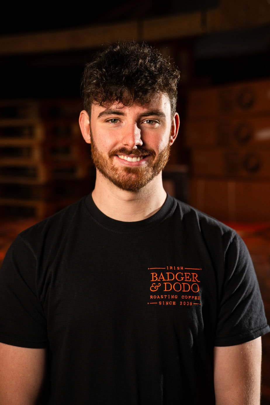 Badger & Dodo. Meet The Team: A young man with curly hair and a beard smiles, wearing a black Badger & Dodo coffee t-shirt.