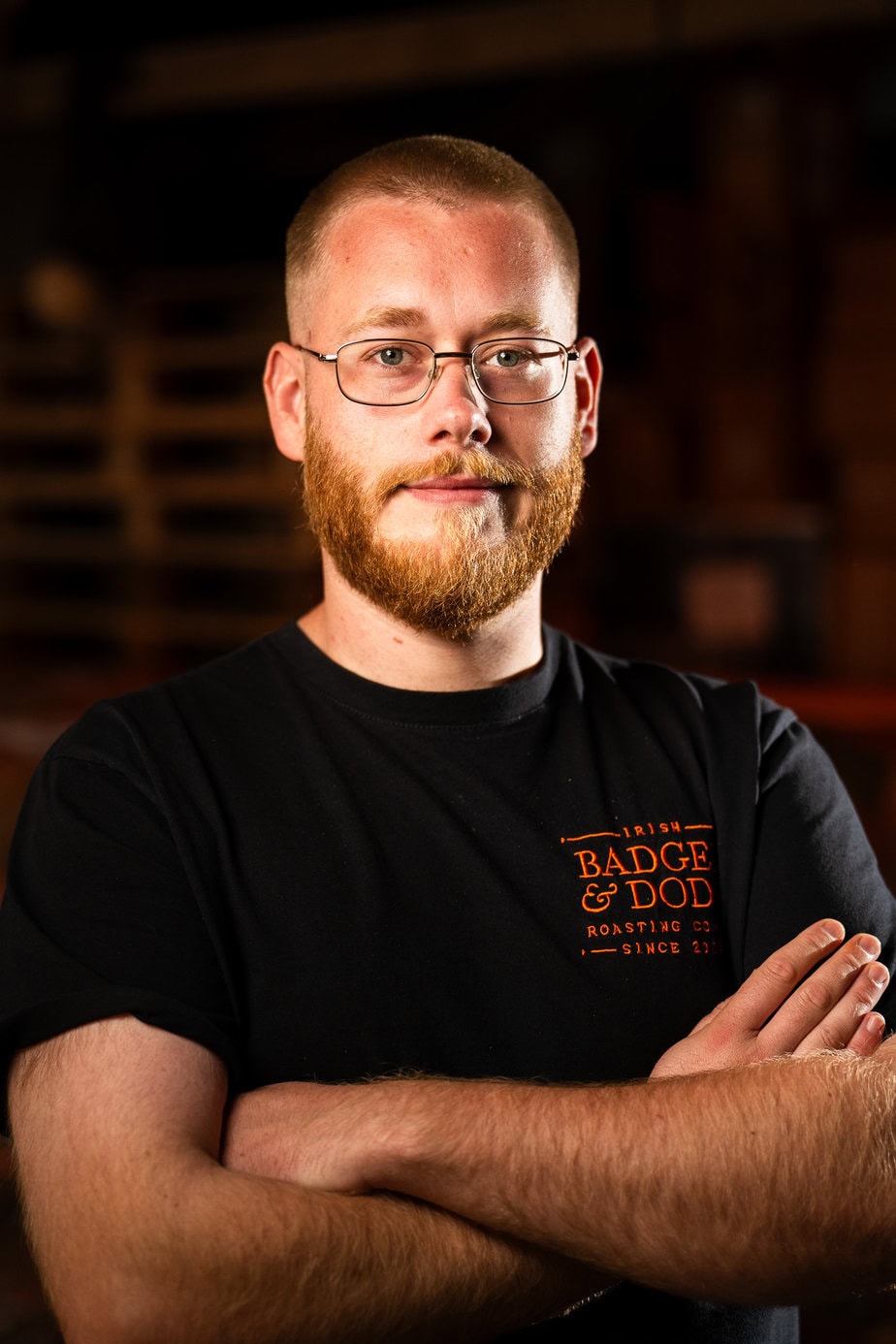 Badger and Dodo Coffee Roasters: A man with glasses, a beard, and a shaved head stands confidently with arms crossed. Hes wearing a black Badge & Dod T-shirt, embodying true team spirit. The dark background features wooden elements, setting the perfect stage to meet the team.
