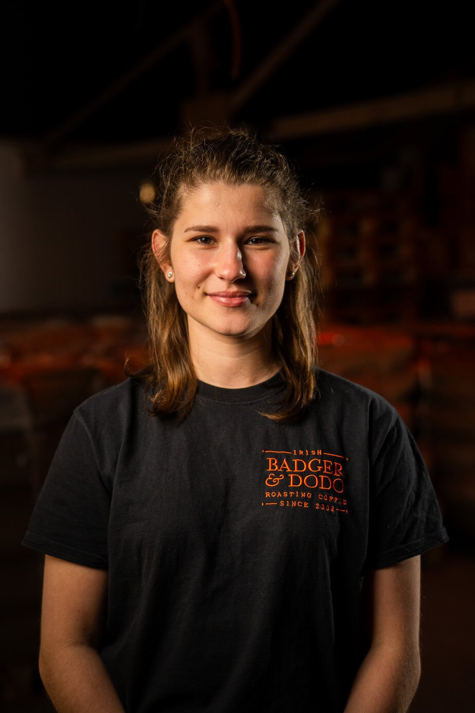 Badger & Dodo. Meet The Team: A woman in a black Badger & Dodo t-shirt smiles in a warmly lit indoor setting.