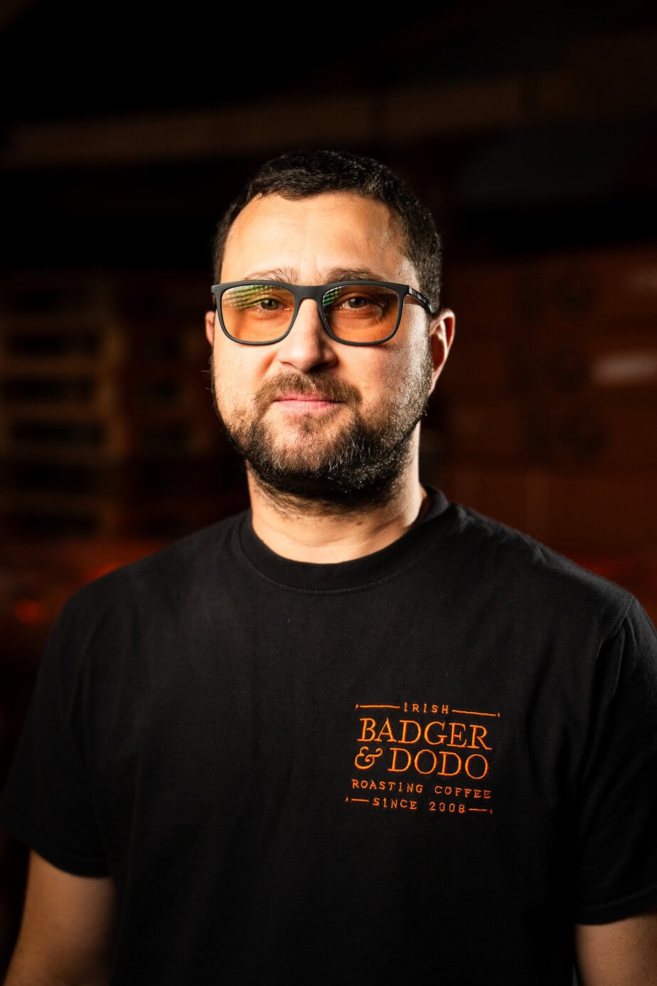 Badger & Dodo. Meet The Team: Man with a beard wearing glasses and a black Badger & Dodo Roasting Coffee t-shirt, standing indoors.