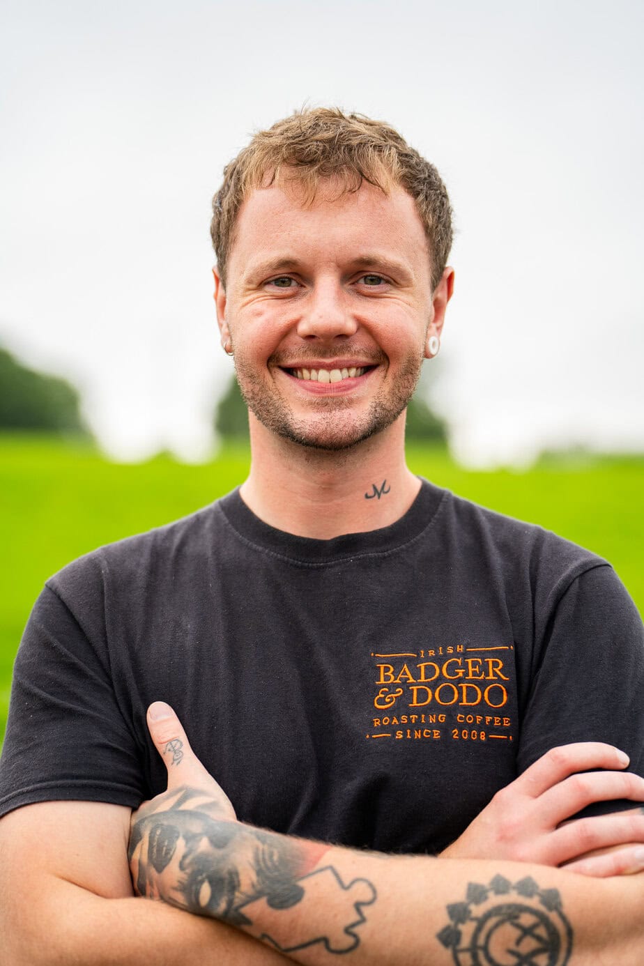 Badger & Dodo. Smiling man with tattoos crosses arms, wearing a black Badger & Dodo t-shirt, standing outdoors—meet the team’s boldest member.