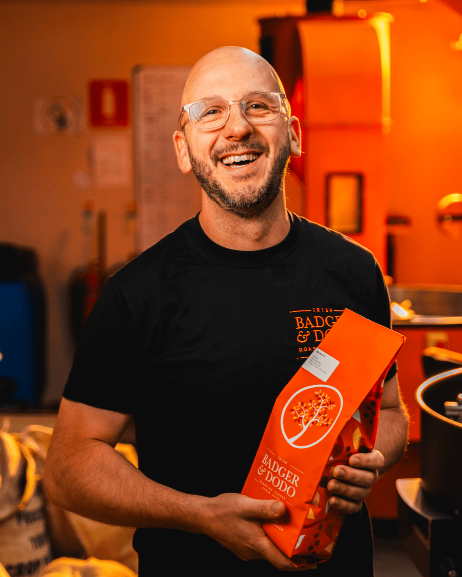 Badger & Dodo. Smiling man with glasses holds a bright orange Badger & Dodo coffee bag in a warm, indoor setting—proudly showcasing the wholesale partner experience.