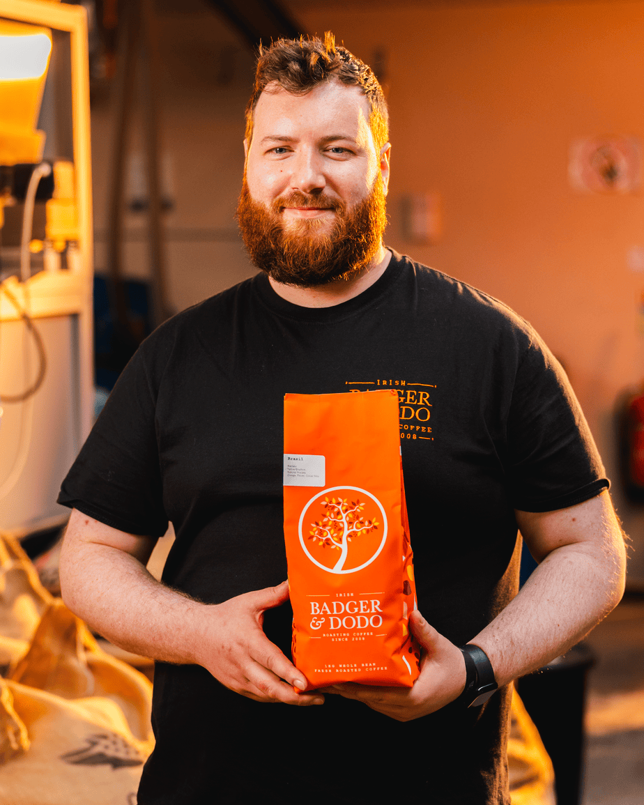 Badger & Dodo. Bearded man in a black shirt holding a bright orange bag of Badger & Dodo coffee, smiling at the camera—perfect for any wholesale partner seeking top-quality beans.