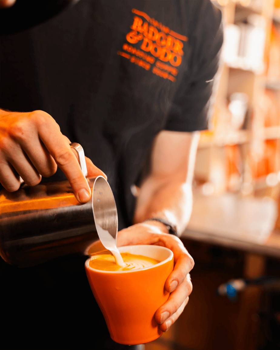 Badger & Dodo. A barista pours steamed milk into a cup of coffee, creating latte art in an orange mug for a wholesale partner.
