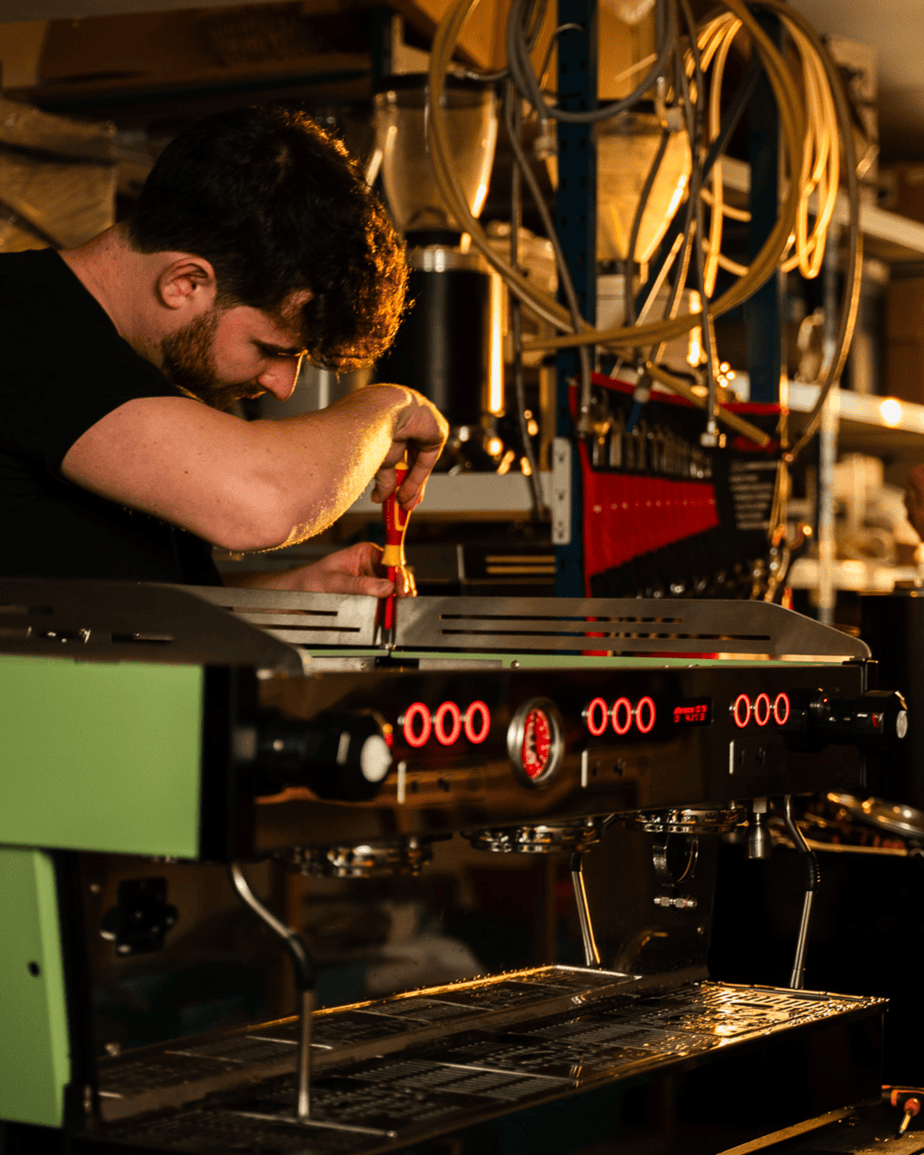 Badger & Dodo. A man, acting as a wholesale partner, uses a screwdriver to repair a coffee machine in a dimly lit workshop.
