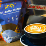 Badger & Dodo. A latte with art in a blue cup sits beside a 400g pack of Juspy Collagen+Protein Powder and a La Marzocco machine.