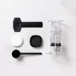 Badger & Dodo. The Aeropress Coffee Maker-Clear Colours kit is disassembled, showing its filter, scoop, stirrer, and cap in transparent hues on a white surface.