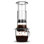 Badger & Dodo. The Aeropress Coffee Maker-Clear Colours presses rich, dark coffee into a glass mug, with vibrant colors highlighting each drip below.