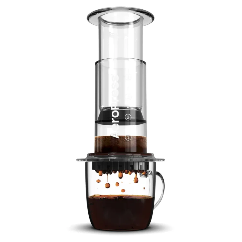 Badger & Dodo. The Aeropress Coffee Maker-Clear Colours presses rich, dark coffee into a glass mug, with vibrant colors highlighting each drip below.