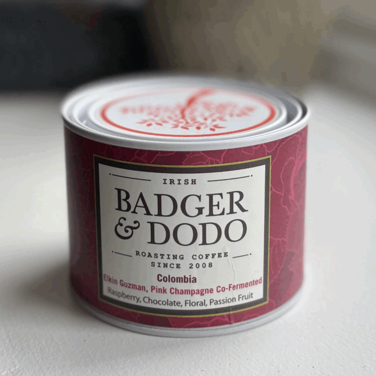 A cylindrical tin of Badger & Dodo coffee with a deep magenta label. The label reads "Irish Badger & Dodo Roasting Coffee Since 2008" and describes the contents as "Colombia – Elkin Guzman, Pink Champagne Co-Fermented" with flavor notes of raspberry, chocolate, floral, and passion fruit. The tin features a decorative lid with a red tree design.