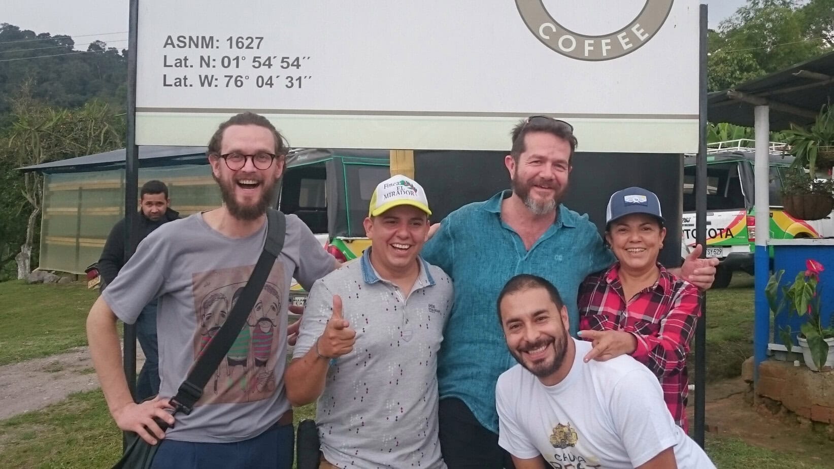 A group of five smiling people pose in front of a large coffee sign that includes geographic coordinates (Lat. N: 01° 54′ 54″, Lat. W: 76° 04′ 31″) and the text "ASNM: 1627". The individuals appear to be enjoying a visit to a coffee farm or cooperative, with one person wearing a cap labeled "Finca El Mirador". A sixth person stands in the background. The setting is outdoors with lush greenery, a blue building, and parked vehicles visible
