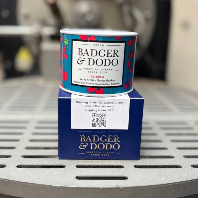 Alt text: A colorful tin of Badger & Dodo coffee with a cherry pattern sits on top of a blue branded box on a metal surface. The label reads “Irish Badger & Dodo Roasting Coffee Since 2008,” with details “Colombia Jairo Arcila – Santa Monica” and flavor notes including maraschino cherry, cola bottles, and amaretto. A sticker on the box shows cupping notes, a score of 88.5, and a QR code.