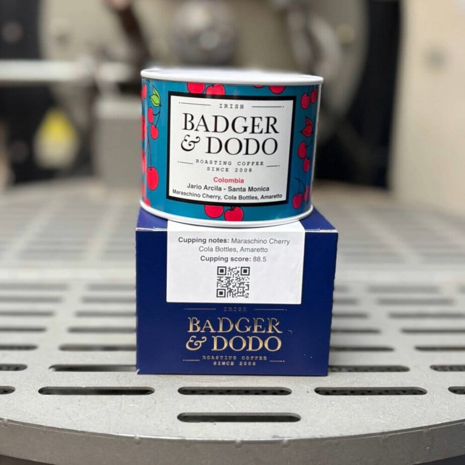 Alt text: A colorful tin of Badger & Dodo coffee with a cherry pattern sits on top of a blue branded box on a metal surface. The label reads “Irish Badger & Dodo Roasting Coffee Since 2008,” with details “Colombia Jairo Arcila – Santa Monica” and flavor notes including maraschino cherry, cola bottles, and amaretto. A sticker on the box shows cupping notes, a score of 88.5, and a QR code.