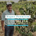 A man wearing a cap and plaid shirt stands in a coffee farm surrounded by green coffee plants. Overlaid text reads “Super Premium Colombia Jairo Arcila.”